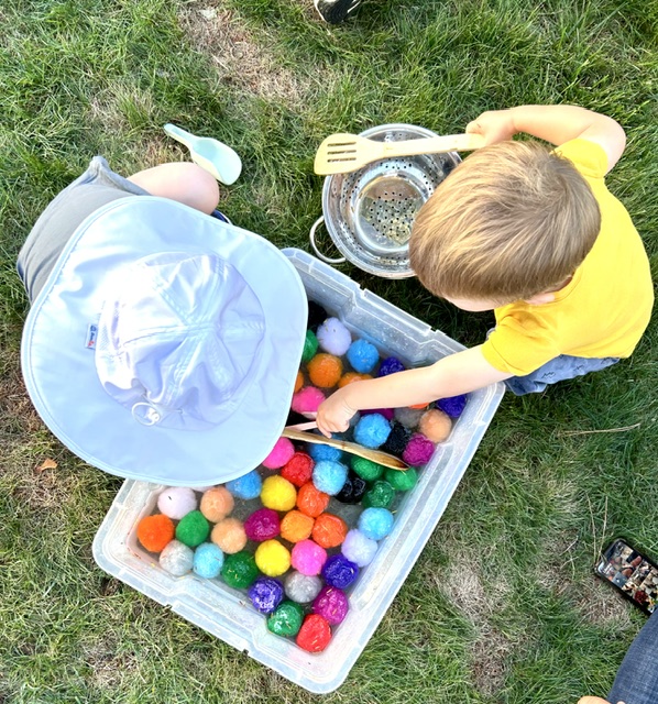 Child scooping colorful pom poms from a sensory bin