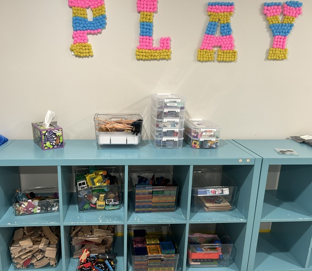PLAY pom-pom wall art above labeled toy shelving